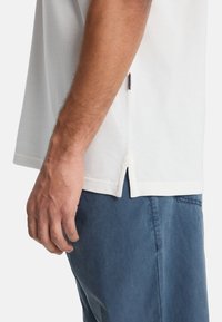 Λευκό πουκάμισο με κοντά μανίκια από υφασμάτινη υφή με πλαϊνές σχισμές, συνδυασμένο με μπλε παντελόνι. Ορατό χέρι ακουμπάει στο ύφασμα.