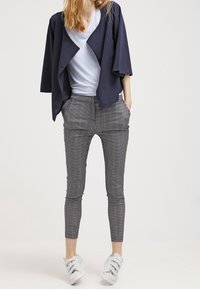 Kvinna iklädd en marinblå öppen jacka, vit V-ringad skjorta, svart- och vitrutiga slim fit byxor och vita kardborreskor, stående med händerna i fickorna.