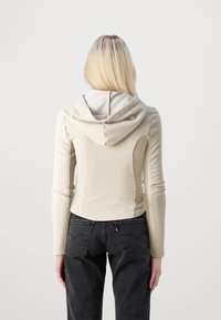En blond person som bär en figurnära beige hoodie och högmidjade svarta jeans, står vänd bort från kameran mot en ljus, enkel bakgrund.