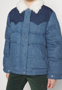Personne portant une veste rembourrée bleue avec un col en polaire blanc et un pantalon vert foncé, se tenant devant un fond uni.