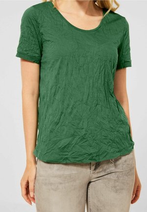 Grünes, zerknittertes Baumwoll-T-Shirt mit kurzen Ärmeln und Rundhalsausschnitt, kombiniert mit hellfarbigen Jeans, die ein dekoratives Reißverschlussdetail aufweisen.