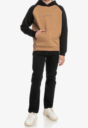 Brauner und schwarzer Hoodie mit Kängurutasche und Raglanärmeln, kombiniert mit schwarzen Hosen und weißen Sneakers, stehend auf weißem Hintergrund.