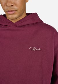 Burgundy hoodie made of cotton with a soft texture; features a large hood and a white embroidered "Pegador" logo on the chest.