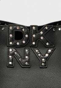 Čierna kožená taška s textúrou, ozdobená veľkými cvočkami a výrazne zobrazenými písmenami "DK NY" z cvočkovaných akcentov.