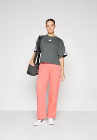 Γκρι κοντομάνικο μπλουζάκι με άσπρο λογότυπο, κοραλλί φόρμες και λευκά αθλητικά παπούτσια. Το μοντέλο κρατά μια μαύρη τσάντα χεριού. Απλός, χαλαρός σχεδιασμός.