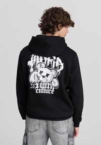 Felpa nera con cappuccio caratterizzata da una grande grafica bianca sul retro, che raffigura un orso con tratti esagerati e il testo "Hybrid Culture" in carattere stilizzato.