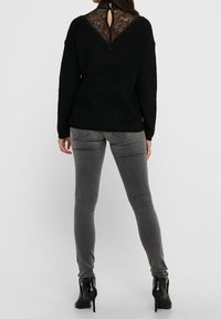 Pull noir en maille avec un détail en dentelle dans le dos et une fermeture à bouton clé, associé à un jean gris ajusté et des chaussures à talons hauts noires.