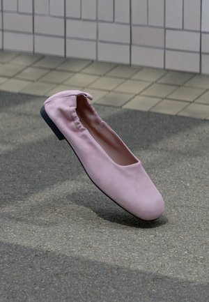 Pojedynczy różowy baletowy balerinek z elastycznym tyłem i okrągłym noskiem unoszący się nad szarym asfaltem w pobliżu ściany wyłożonej kafelkami.