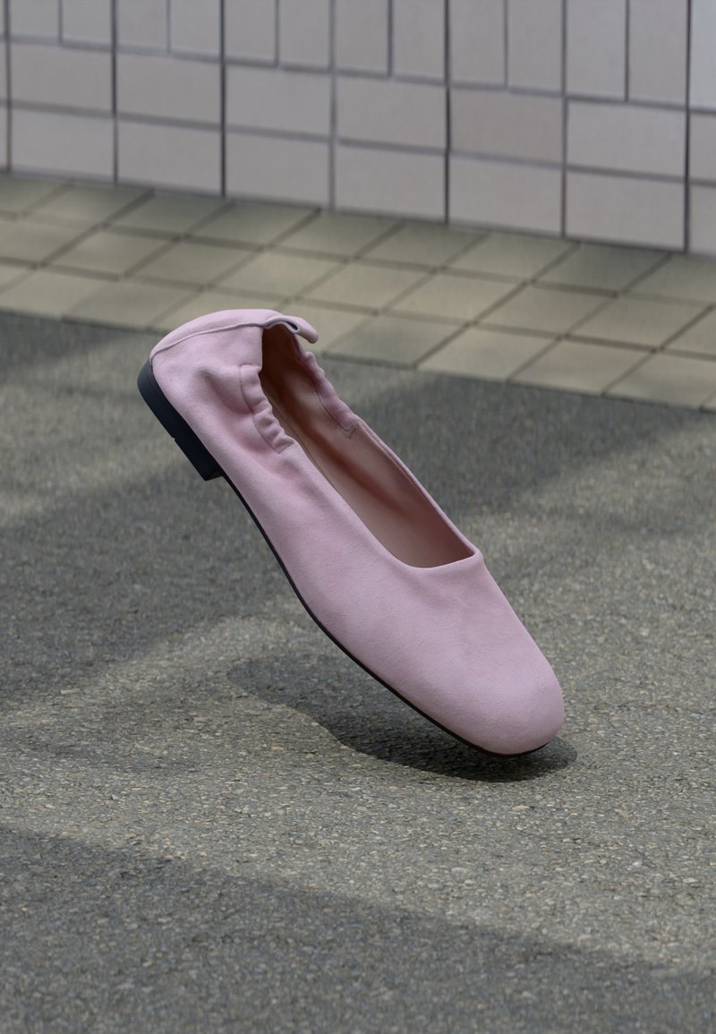 Une ballerine plate rose unique avec un dos élastiqué et un bout arrondi flottant au-dessus d’un bitume gris près d’un mur carrelé.