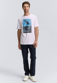 Hvid bomuld T-shirt med et blåt grafisk print, der viser en helikopter over vandet. Kombineret med mørke bukser og hvide sneakers.