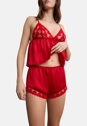 Donna che indossa un completo intimo con canottiera e pantaloncini in pizzo e raso rossi, solleva l'orlo della canottiera per mostrare l'addome.