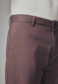 Brune bomuldsbukser med en glat tekstur, der har en standard linning og sidelommer. Simpelt design med lukning ved én enkelt knap.