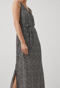 Femme portant une robe maxi sans manches noire avec un imprimé floral blanc et une fente sur le côté, se tenant contre un fond clair.