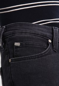 Vaqueros de mezclilla negros con una textura suave, que presentan un pequeño logo plateado de "Lee" en el bolsillo delantero y detalles de costura clásica.
