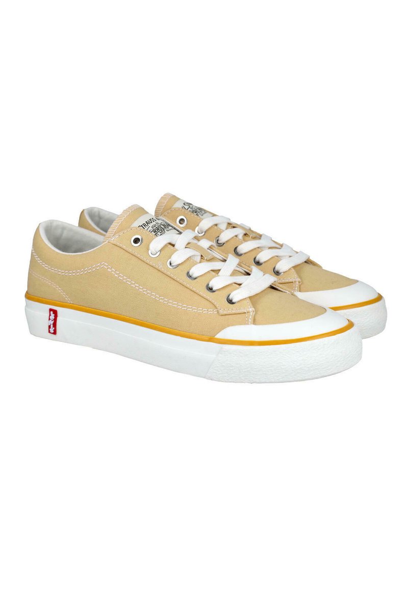 Zapatillas bajas de lona beige con suelas de goma blancas, cordones blancos, ribete amarillo y etiqueta de logo roja en el lateral.