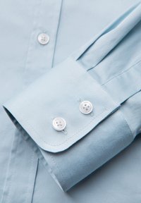 Poignet de chemise bleu clair avec deux boutons blancs et couture visible, reposant sur le panneau avant boutonné de la chemise.