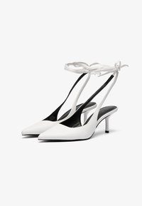 Valkoiset nahkaiset slingback-kengät, joissa on terävä kärki, ohuet mustat korot ja säädettävä valkoinen nauha nilkan ympärillä. Silkkinen pinta, minimalistinen muotoilu.