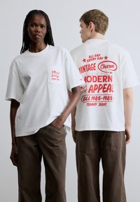 Camisetas blancas de manga corta con estampados gráficos rojos en el bolsillo delantero y en la espalda. El material parece suave; corte holgado; combinadas con pantalones marrones.