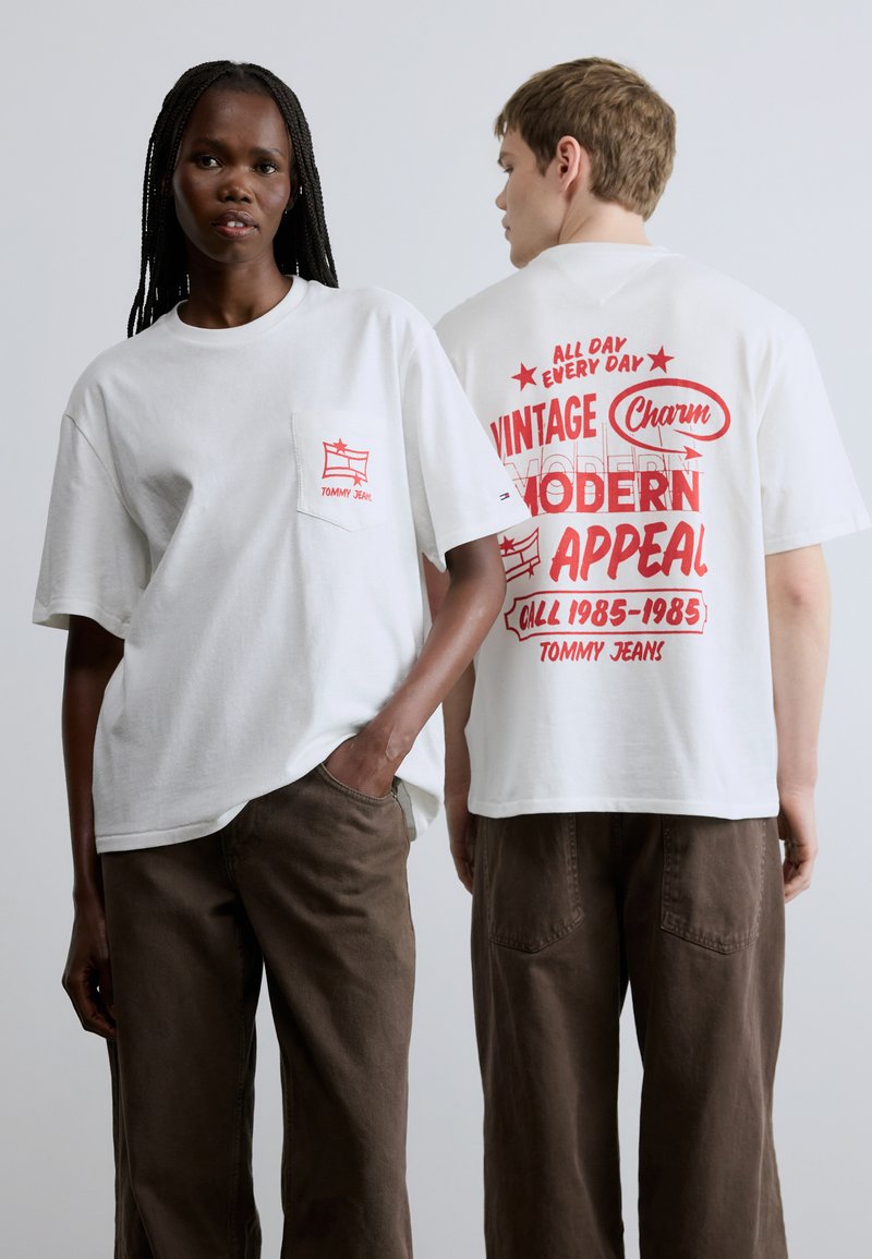 Camisetas blancas de manga corta con estampados gráficos rojos en el bolsillo delantero y en la espalda. El material parece suave; corte holgado; combinadas con pantalones marrones.