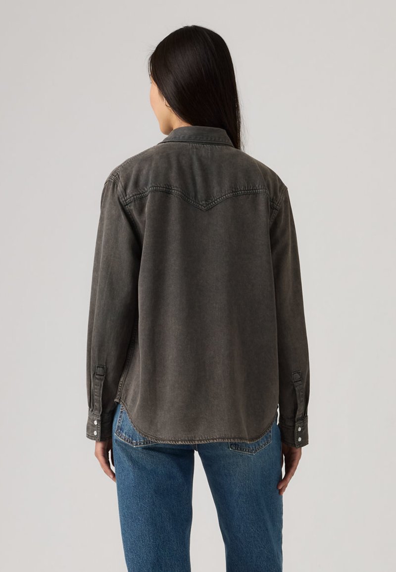 Woman with long dark hair wearing a faded black long-sleeve button-up shirt and blue jeans, viewed from the back against a plain background.