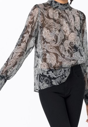 Kobieta ubrana w przejrzystą czarną bluzkę z białymi wzorami paisley, z wysokim kołnierzem i długimi rękawami, zestawioną z czarnymi spodniami.