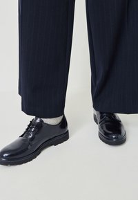 Čierne kožené topánky s lesklým povrchom, šnurovací dizajn a hrubá podrážka. Nosené s šedými ponožkami a námorníckymi pruhovanými nohavicami.