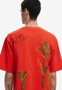 Rød bomulds t-shirt med en afslappet pasform, prydet med grønne og gule blomsterbroderier og teksten "JEG ELSKER DEN, JEG ER" i sort syning.