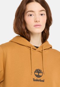 Hnědá mikina s kapucí, která má šňůrku na stažení, žebrované manžety a černé logo Timberland se symbolem stromu na hrudi.