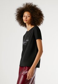 Mladá žena s kučeravými vlasmi, oblečená v čiernom tričku s potlačou a lesklou tmavočervenou koženou sukňou, stojaca pred jednofarebným pozadím.