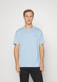 Hellblaues Baumwoll-T-Shirt mit rundem Halsausschnitt und kurzen Ärmeln. Mit kleinem Logo auf der Brust. Kombiniert mit schwarzen Hosen.