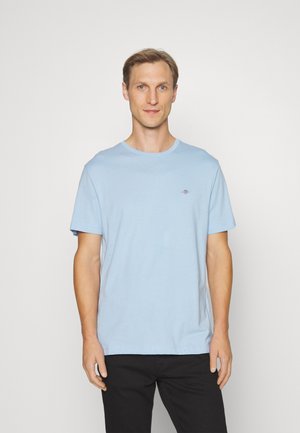 Hellblaues Baumwoll-T-Shirt mit rundem Halsausschnitt und kurzen Ärmeln. Mit kleinem Logo auf der Brust. Kombiniert mit schwarzen Hosen.