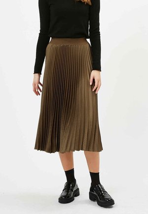 Femme portant une jupe midi plissée marron, un haut noir à manches longues, des chaussettes noires et des mocassins à plateforme noirs, debout sur un fond blanc.