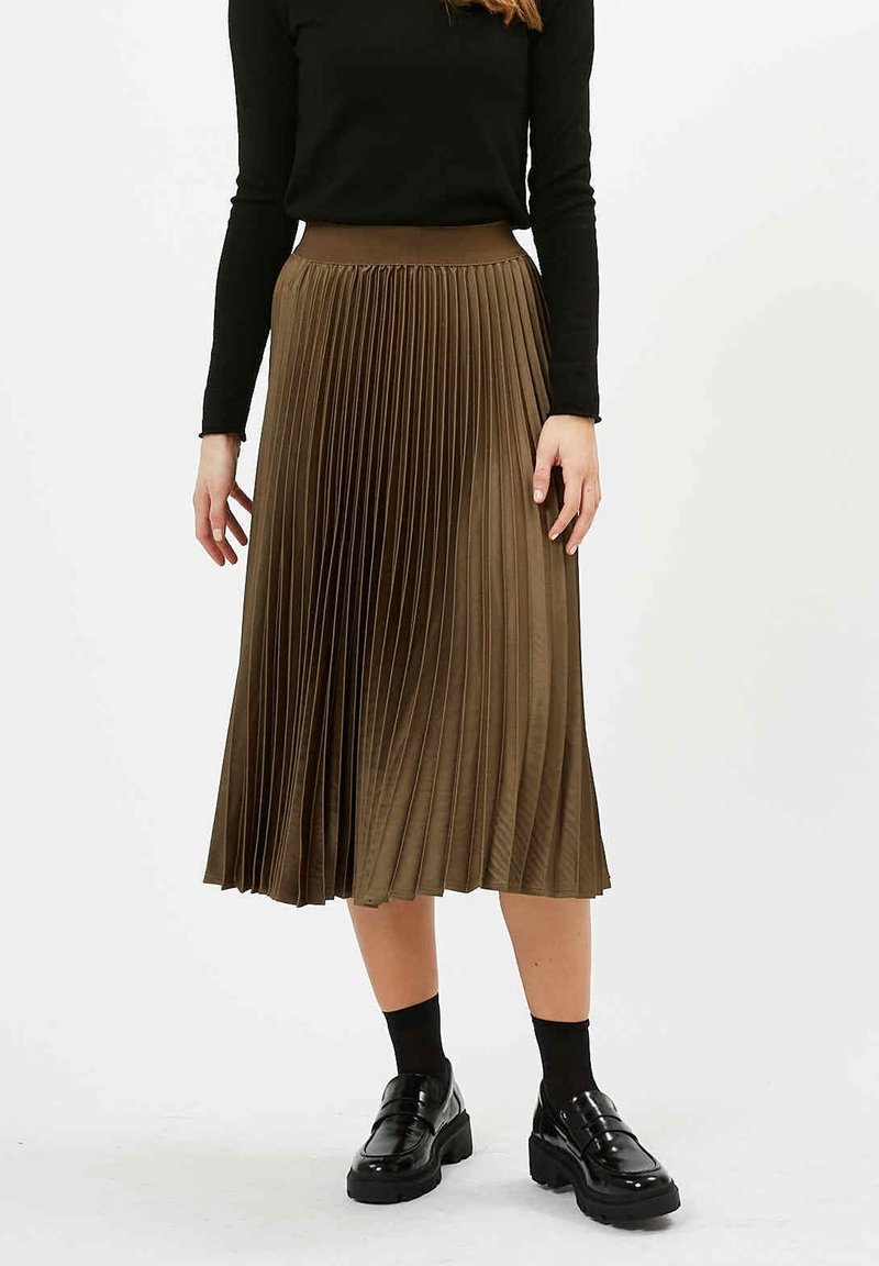 Femme portant une jupe midi plissée marron, un haut noir à manches longues, des chaussettes noires et des mocassins à plateforme noirs, debout sur un fond blanc.
