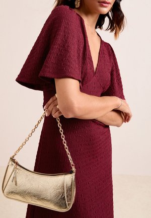 Femme en robe bordeaux texturée avec manches papillon tenant un petit sac à main doré avec une bandoulière chaîne sur un fond neutre.