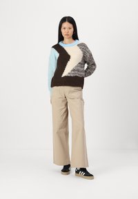 Wielokolorowy sweter dzianinowy z geometrycznym wzorem w kolorze brązowym, jasnoniebieskim, kremowym i szarym; zestawiony z beżowymi spodniami typu wide-leg i czarnymi sneakersami.