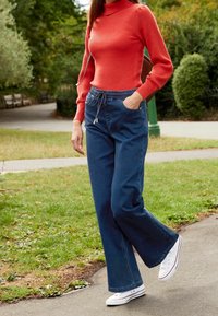 Pull à col roulé rouge côtelé, pantalon en denim bleu à jambes larges avec taille à cordon, et baskets blanches sur un chemin herbeux en plein air.