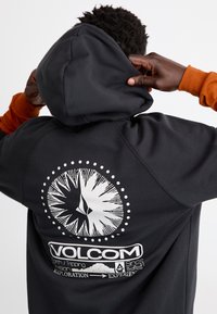 Fekete kapucnis pulóver, nagy fehér iránytű grafikával és márkalogóval a hátsó részen; húzózsinóros kapucnival és bordás mandzsettákkal rendelkezik.