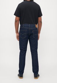 Tmavě modré džínové kalhoty s rovně střiženým designem, s kapsami na zadní straně a koženou nášivkou na pasu. Nahoře černé tričko.