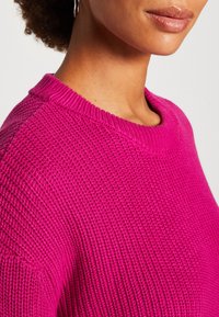 Gros plan sur une personne portant un pull tricoté texturé rose vif avec un col rond, montrant le cou et une partie du bas du visage.