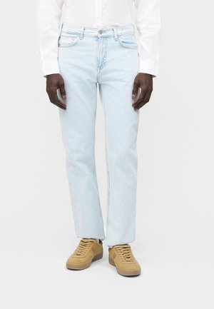 Mees kannab helesiniseid sirge säärega teksapükse, valget nööpidega särki ja beeže seemisnahast vabaajajalatsit, seisab ühevärvilise tausta ees.