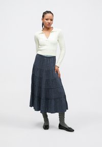 Młoda kobieta z warkoczami, ubrana w biały dziany sweter, niebieską midi spódnicę w kratę, szare ocieplacze na nogi oraz czarne baleriny.