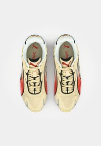 Béžové sportovní tenisky s černými a červenými akcenty, texturovaný síťovaný materiál a polstrovaná podrážka. Logo 'Puma' viditelné na jazyku.