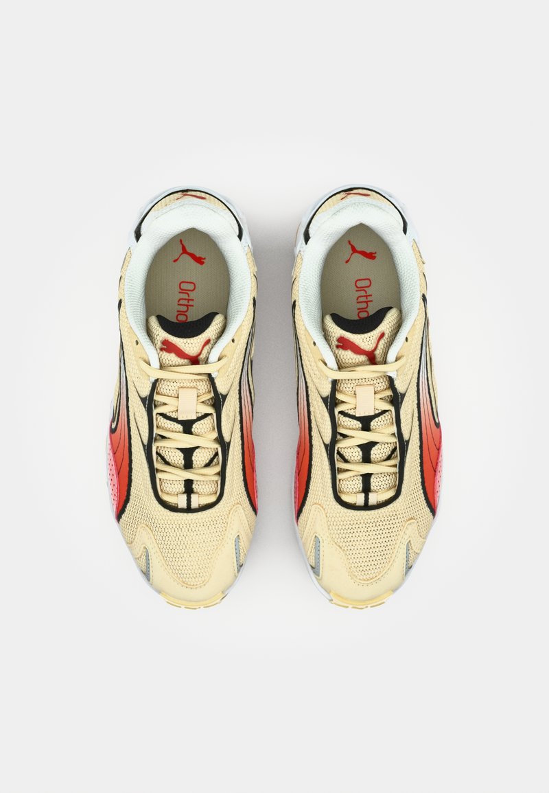 Baskets de sport beiges avec des accents noirs et rouges, en matériau maillé texturé, et une semelle rembourrée. Logo 'Puma' visible sur la languette.