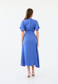 Niebieska lniana sukienka z falbaniastymi rękawami, dopasowaną talią i zwiewnym dołem. Linia hemu jest asymetryczna. Założona z beżowymi sandałami na obcasie.