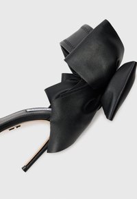 Black high-heeled sandal with a large, textured bow detail. Features a sleek strap design and a thin stiletto heel. Smooth leather material.