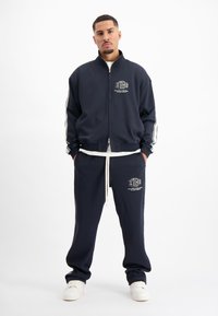 Marine trainingspak met witte strepen langs de mouwen en benen, ritsjack, elastische tailleband, voorzakken en logo detail op de kleding.