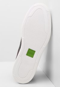 Braune Sneaker mit weißer Gummisohle, strukturiertem Profil und grünem Akzentlabel. Verfügt über Metallösen für die Schnürsenkel im Obermaterial.