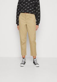 Pantaloni beige a pantaloni corti con gamba dritta, realizzati in twill di cotone, abbinati a una felpa nera con testo bianco ripetuto e sneakers nere.