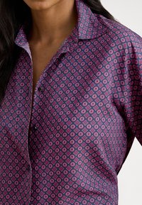 Lilla mønstret skjorte med geometriske design i hvitt og rosa. Har klassisk krage og knappelukking foran. Glatt tekstur.