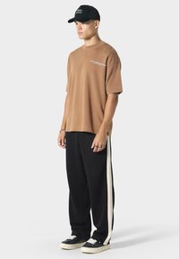 Brun oversized T-shirt med korte ærmer og logo-detalje, parret med sorte afslappede bukser med en hvid sidestribe og sneakers.